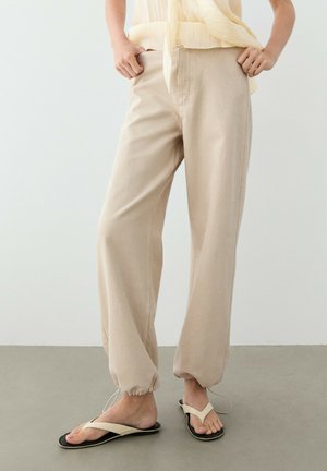 Pantalon beige ample avec poignets élastiques porté avec des sandales crème à lanières, assorti à un haut sans manches jaune clair, debout sur un sol gris.