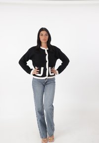 Cárdigan negro con ribete blanco, con un frente abotonado y dos bolsillos decorativos, combinado con jeans azul claro y sandalias transparentes.