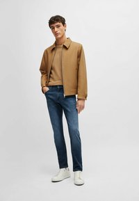 Veste zippée beige, pull beige, jeans bleu foncé et baskets blanches. Textures lisses et coupe moderne, avec un col et des poches latérales.