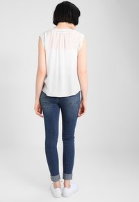Top bianco senza maniche con schiena arricciata, abbinato a jeans in denim aderenti, blu scuro con risvolto sulla caviglia, e sneakers bianche.