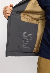 Chaqueta exterior gris con un forro interior beige, que presenta un bolsillo y etiquetas de materiales. Incluye detalles sobre resistencia al agua y aislamiento.