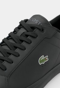 Baskets en cuir noir avec lacets noirs, logo crocodile vert brodé sur le côté et nom de la marque embossé sur la languette.