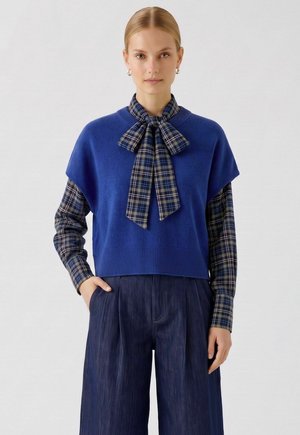 Frau trägt blauen ärmellosen Pullover über karierter Hemd mit Schleife und weit geschnittene dunkle Denim-Hose, steht mit einer Hand in der Tasche.