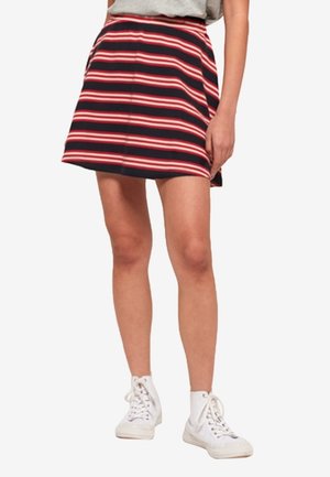 Femme portant une jupe bleu marine à rayures horizontales rouges et blanches, un haut gris et des baskets montantes blanches, debout avec une main sur le côté.