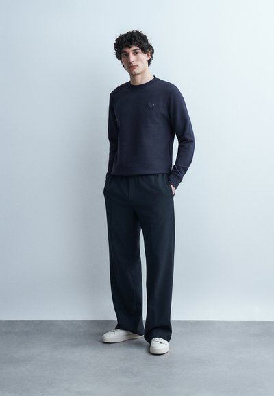 Νέος άνδρας με σγουρά μαλλιά, φοράει μπλε ναυτικό φούτερ και μαύρο παντελόνι με φαρδύ πόδι, στέκεται με τα χέρια στις τσέπες μπροστά σε απλό φόντο.