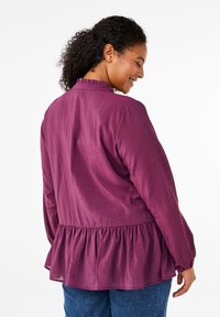 Blusa lunga viola con maniche a sbuffo, caratterizzata da un colletto a volant e un orlo peplum. Realizzata in un tessuto testurizzato, con una vestibilità rilassata e una silhouette morbida.