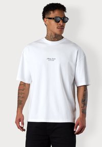 Weißes Baumwoll-T-Shirt mit lockerer Passform, rundem Ausschnitt und kurzen Ärmeln. Mit einem kleinen „Esteem“-Logo in schwarzer Stickerei auf der Brust.