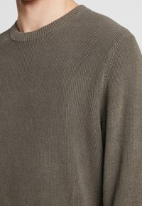 Khakifarbenen Pullover mit gerippter Textur und Rundhalsausschnitt. Lange Ärmel, weiches Material. Besitzt ein dezentes, gleichmäßiges Strickmuster.