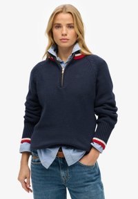 Marineblauwe trui met een kwartrits, voorzien van ribbelmanchetten met rood, wit en blauwe accenten en een gestructureerd breipatroon. Gelaagd over een lichtblauw overhemd.