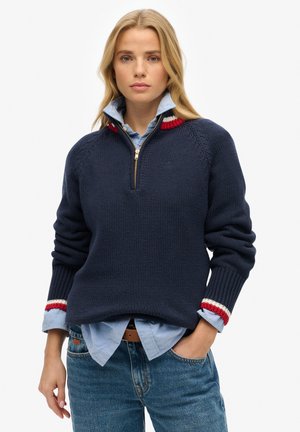 Pull marin avec fermeture à quart, présentant des poignets côtelés avec des accents rouge, blanc et bleu et un design en tricot texturé. Superposé à une chemise bleu clair.