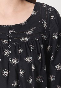 Blouse noire avec petits motifs floraux blancs et pastel, boutons sur le devant, détails en dentelle et tissu froncé sous la poitrine.