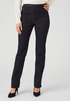 Donna che indossa pantaloni neri aderenti e scarpe nere con tacco alto, in piedi con una mano sul fianco e un top bianco a maniche lunghe.
