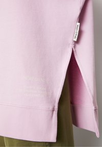 Sudadera de un ligero color morado con dobladillo y abertura lateral, etiqueta blanca "Marc O'Polo", texto tenue en la tela, llevada sobre pantalones verde oliva.
