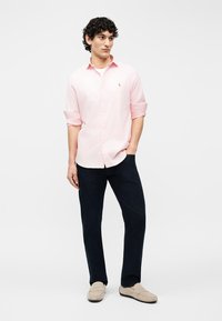 Νέος άνδρας που στέκεται με το ένα χέρι στην τσέπη, φοράει ανοιχτό ροζ ριγέ πουκάμισο, σκούρο παντελόνι και μπεζ μοκασίνια, με φόντο λευκό.