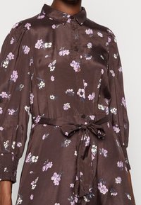Braunes Satin-Kleid mit Kragen, Knopfverschluss vorne und einer Bindegürtel in der Taille. Mit floralen Mustern in Lila und Weiß. Lange, gepuffte Ärmel mit elastischen Bündchen.