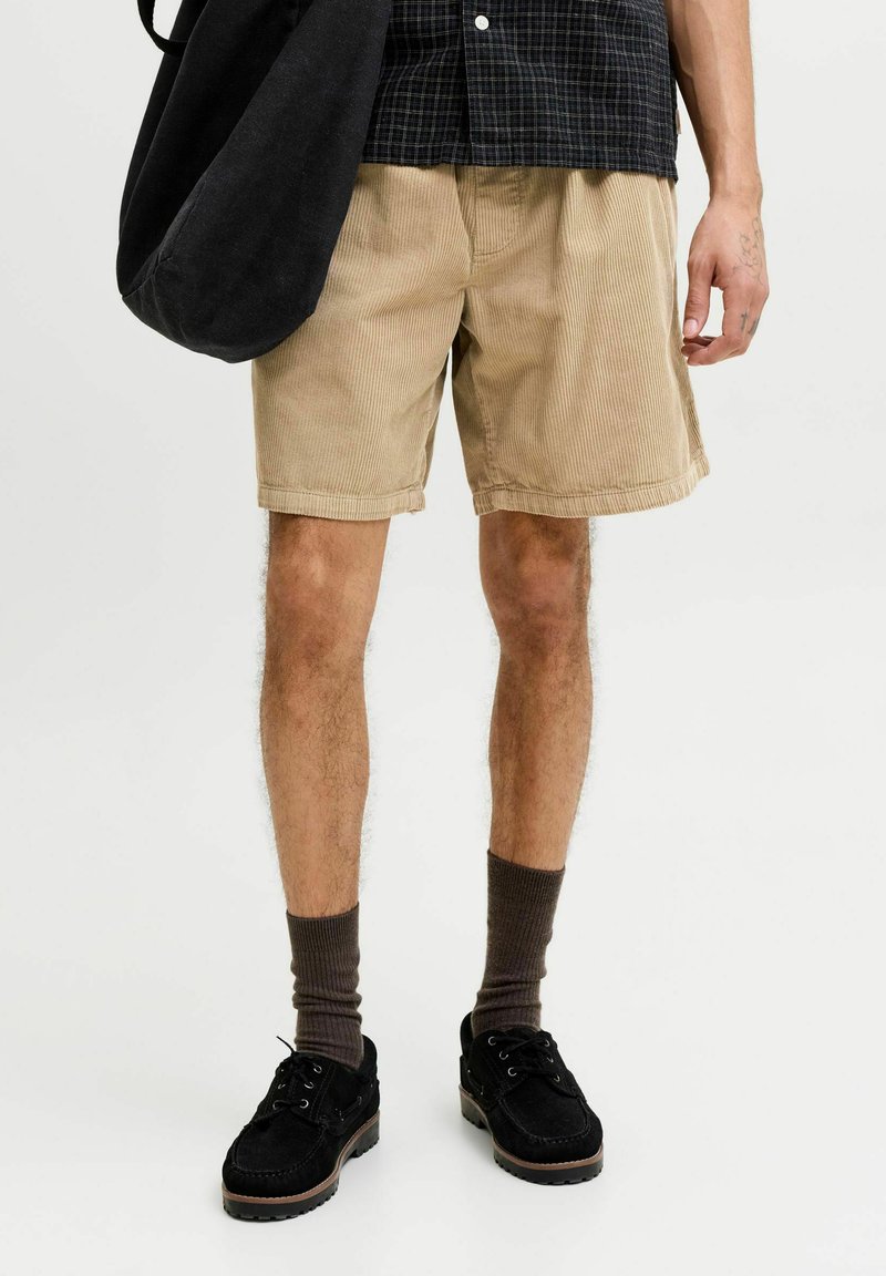 Homme portant un short en velours côtelé beige, des chaussettes foncées, des chaussures noires, tenant un grand sac noir, avec une chemise noire à carreaux.