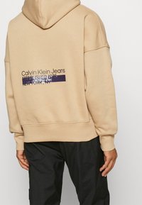 Sweatshirt beige à capuche avec le texte "Calvin Klein Jeans". Présente une coupe décontractée, des poignets et un ourlet côtelés. Design simple avec un détail de logo imprimé.
