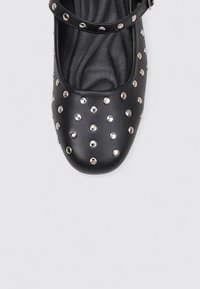 Zapato de cuero negro con punta redondeada, adornado con tachuelas plateadas en la parte superior y una correa ajustable en el tobillo. Plantilla texturizada visible.