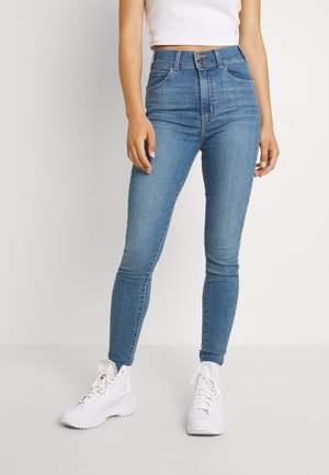 Femme portant un jean skinny bleu taille haute avec fermeture à double bouton et des baskets blanches épaisses sur un fond clair uni.