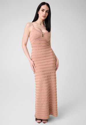 Frau mit langen dunklen Haaren, die ein ärmelloses, pink-gold gestreiftes bodenlanges Kleid und dunkle offene Schuhe trägt, steht vor einem schlichten Hintergrund.