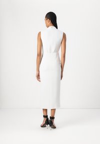 White sleeveless dress with a high neckline and back zipper. Shaped fit, midi length, smooth fabric, finished with black heeled sandals.