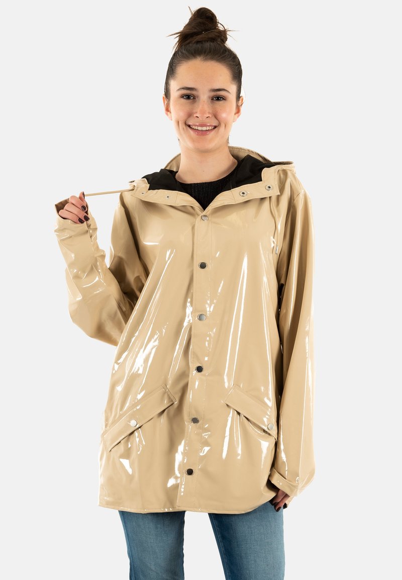 Impermeable brillante beige con capucha, botones de presión, dos bolsillos delanteros y forro interior negro, llevado sobre un suéter y jeans.