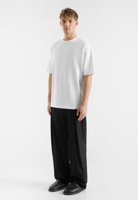 Hvid t-shirt med korte ærmer og rund hals, parret med sorte vide bukser og sorte slip-on sko, sat mod en ensfarvet baggrund.