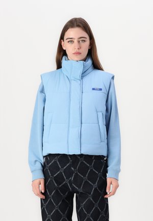 Lichtblauw puffer vest met hoge kraag, voorzakken en een logopatch. Gedragen over een blauw langarmshirt en zwart met patroon broeken.