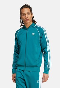 Tyrkysový Adidas trenýrkový komplet s bílými bočními pruhy, vysokým límcem a zipovým uzávěrem. Obsahuje logo Adidas na hrudi a na kalhotách.