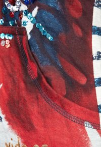 Rødt stof med blå og hvide abstrakte mønstre, smykket med paljetter i rødt, blåt og turkist. Har en rund halsudskæring og syede kanter.