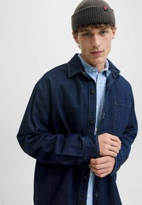 Camicia di jeans blu scuro con chiusura a bottoni, taschino sul petto e polsini regolabili, indossata sopra una camicia a colletto blu chiaro.