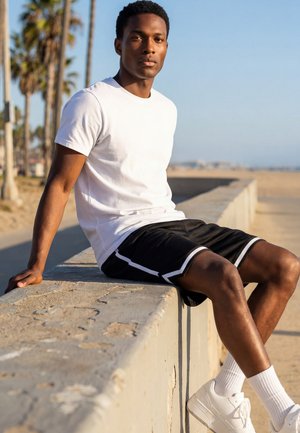 Jeune homme en t-shirt blanc, short noir et baskets blanches assis sur un rebord en pierre près d'une plage avec des palmiers en arrière-plan.
