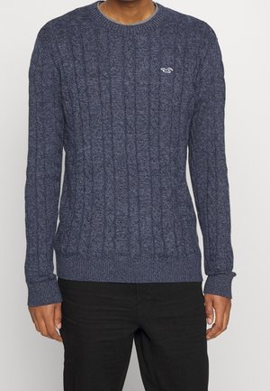 Pull en maille torsadée bleu marine avec un col et des poignets côtelés, arborant un petit logo sur la poitrine, fabriqué à partir d'un tissu doux et texturé.