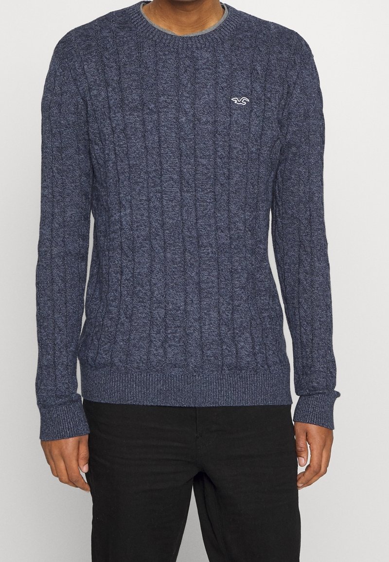 Pull en maille torsadée bleu marine avec un col et des poignets côtelés, arborant un petit logo sur la poitrine, fabriqué à partir d'un tissu doux et texturé.