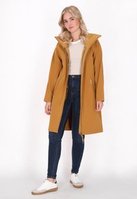 Sinepės geltonumo iki kelių ilgio paltas su dideliu kapišonu ir pilnu užtrauktuku, derinamas su baltu dryžuotu marškinėliu, tamsiomis liesomis džinsais ir baltais sportbačiais.