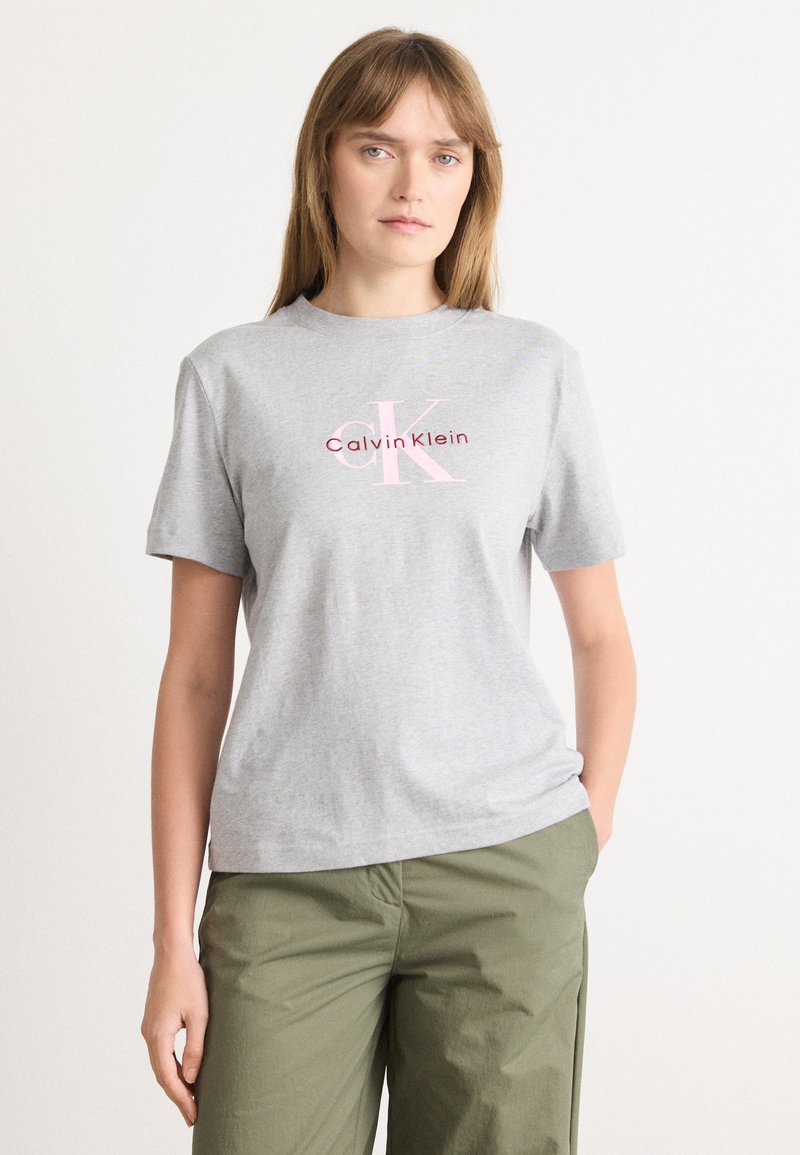 Camiseta de algodón gris con mangas cortas, que presenta un logotipo de Calvin Klein en rosa y rojo impreso en el frente. Corte casual, diseño simple.