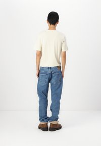 Lichtbeige t-shirt en blauwe denim jeans, met een relaxte pasvorm, zichtbare achterzakken en bruine werklaarzen met zwarte zolen.