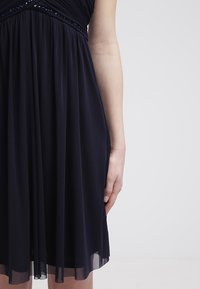Bras droit d'une personne portant une robe noire au genou avec une jupe plissée et une bordure en sequins à la taille, sur un fond gris clair uni.