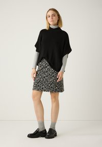 Schwarzes Poncho-Oberteil über einem grauen Langarmhemd, kombiniert mit einem grauen Rock im Leopardenmuster. Schwarze Schuhe und graue Socken runden das Outfit ab.