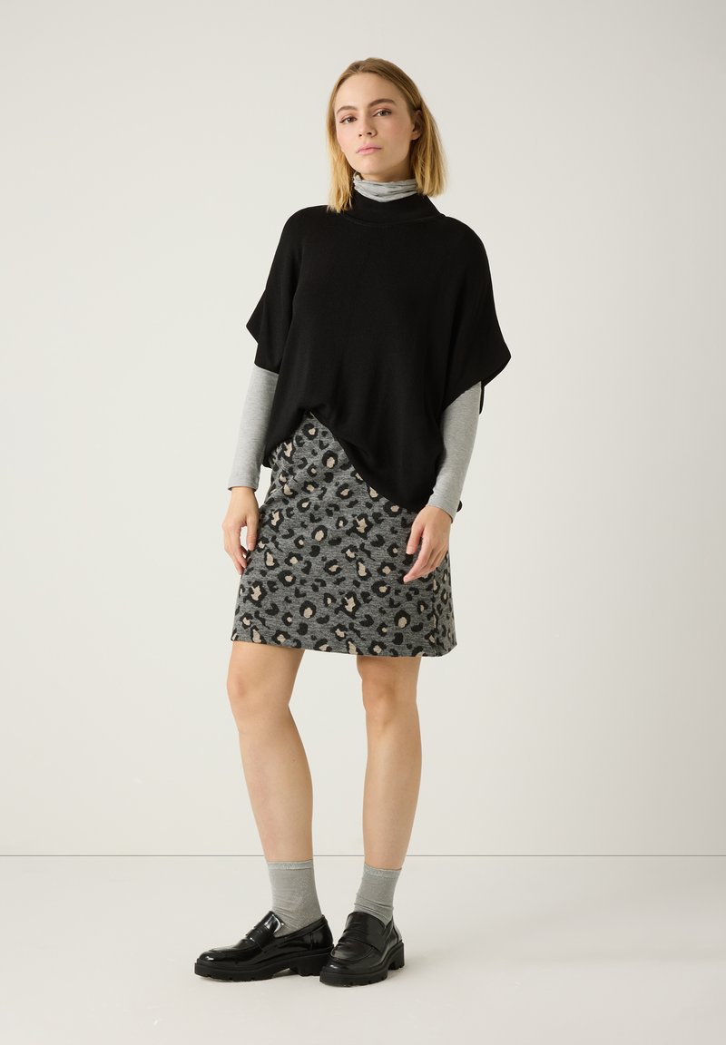 Schwarzes Poncho-Oberteil über einem grauen Langarmhemd, kombiniert mit einem grauen Rock im Leopardenmuster. Schwarze Schuhe und graue Socken runden das Outfit ab.