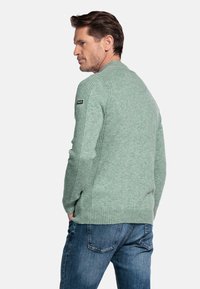 Groene gebreide trui met ribbelstructuur, ronde halslijn en een zwart label op de schouder. Gecombineerd met blauwe spijkerbroek.