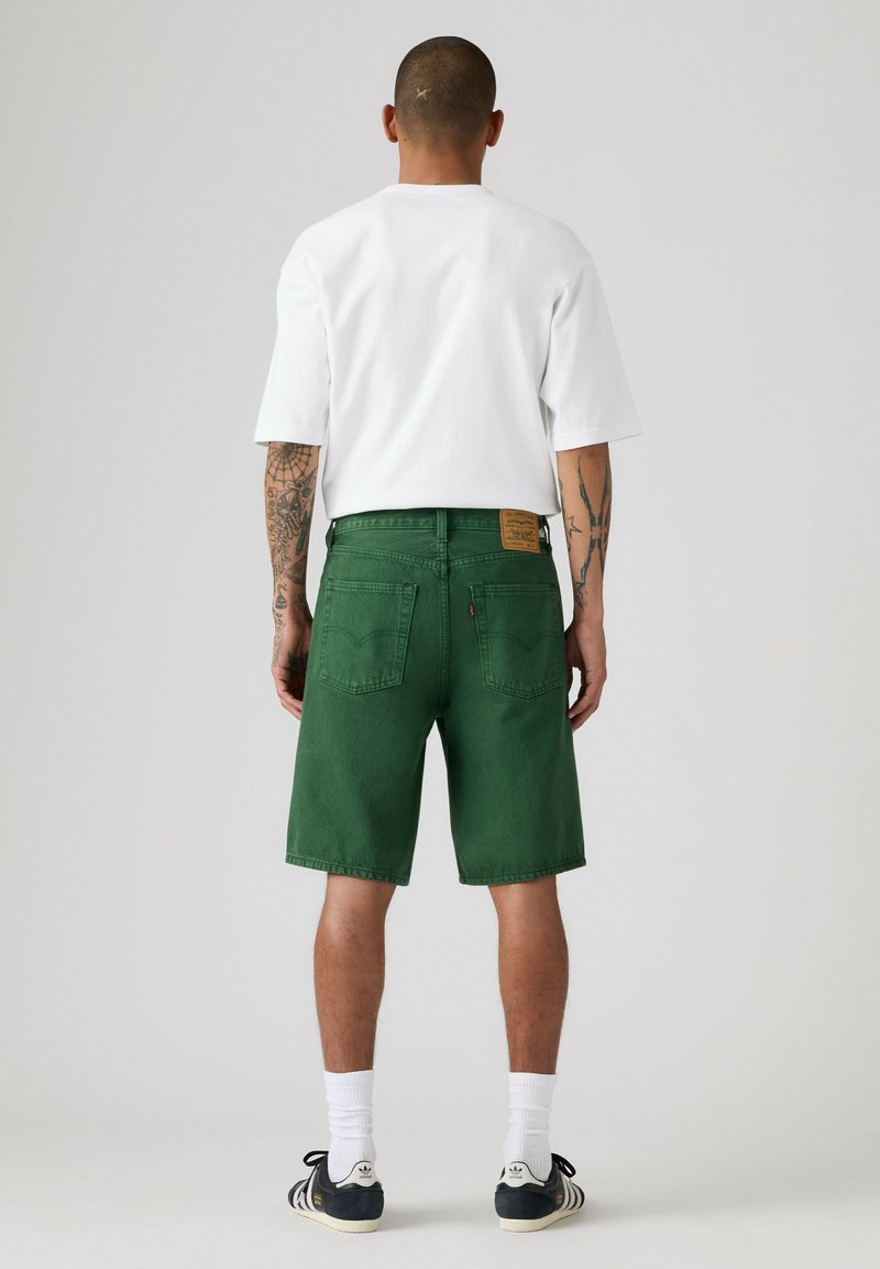 Mann mit Tätowierungen, der ein weißes T-Shirt, grüne knielange Shorts, weiße Socken und schwarze Adidas-Sneakers trägt und mit dem Rücken zum Betrachter auf schlichtem Hintergrund steht.