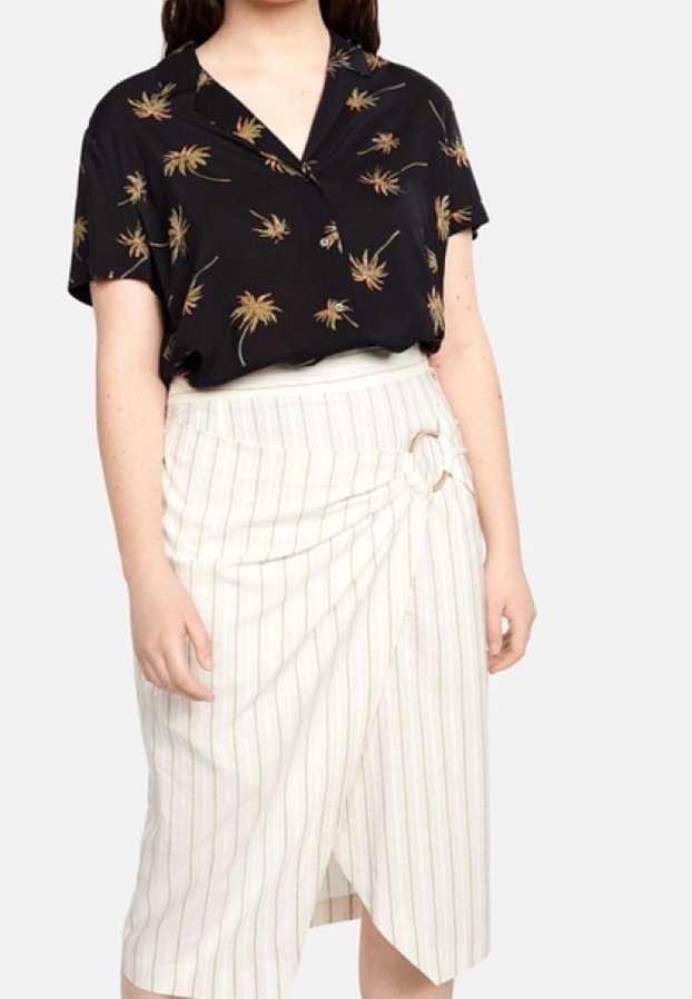 Femme portant une chemise noire à manches courtes avec imprimé floral et une jupe portefeuille blanche à fines rayures avec une boucle latérale.