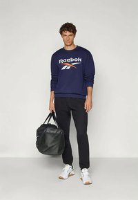 Felpa Reebok blu navy con logo bianco e arancione, abbinata a pantaloni neri, mentre tiene una borsa da palestra nera e indossa scarpe da ginnastica bianche.
