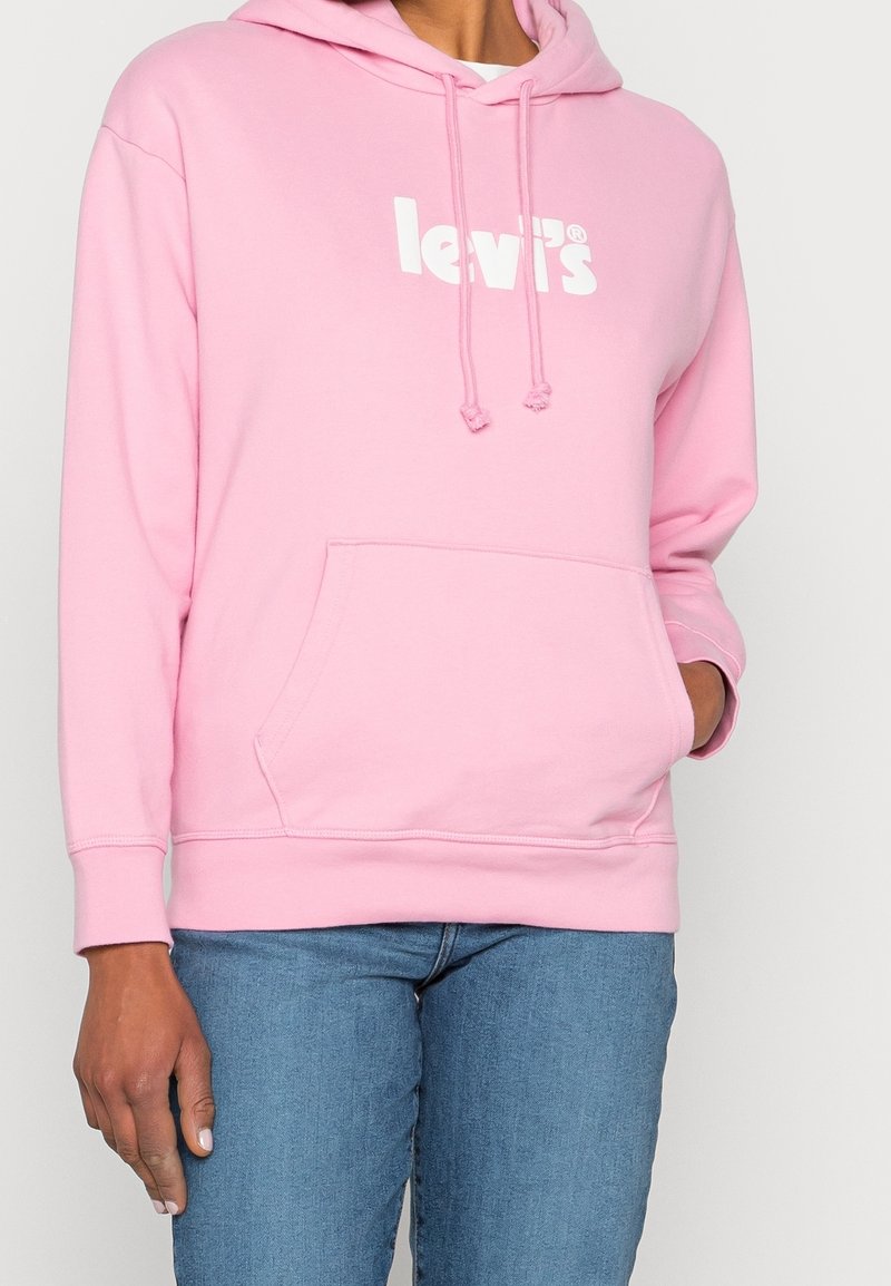 Persona che indossa una felpa chiara rosa di Levi's con logo bianco e jeans blu, una mano in tasca e l'altra al suo fianco.