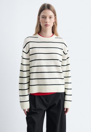 Jeune femme aux longs cheveux blonds portant des boucles d'oreilles créoles dorées, un pull blanc à rayures horizontales noires, un sous-pull rouge et un pantalon noir.