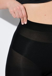 Leggings neri con un ampio cinturino elastico e sezioni trasparenti e decorate. Il tessuto appare liscio e leggermente lucido.