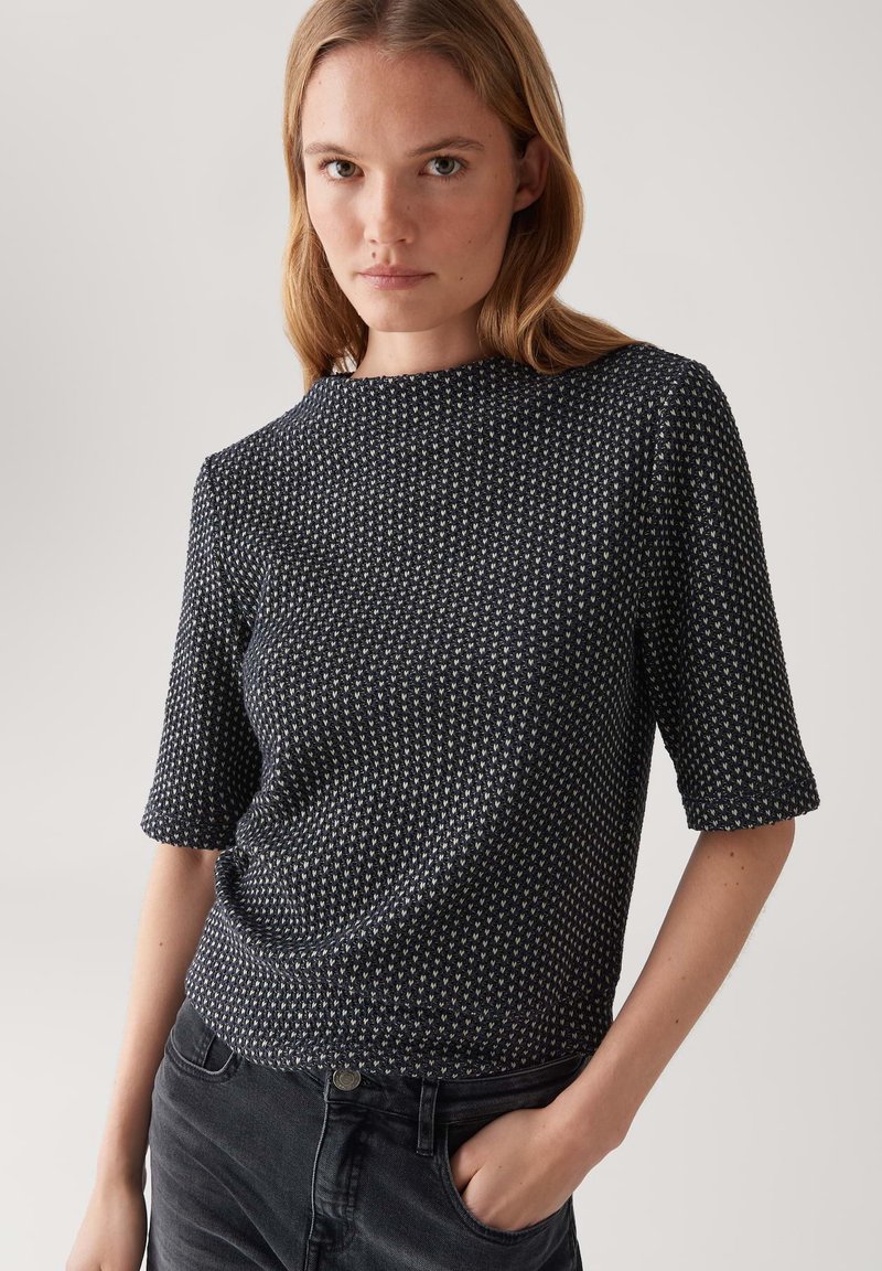Femme aux cheveux châtain clair portant un haut à manches courtes texturé noir et blanc et un jean noir, se tenant avec une main dans la poche.