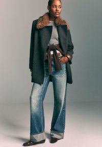 Donna che indossa un cappotto scuro con colletto in pelliccia, maglione grigio, top marrone annodato, jeans larghi con risvolti, ballerine marroni e un braccialetto dorato, in piedi con una mano in tasca.