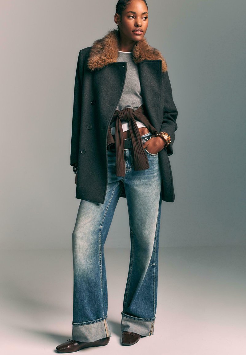 Donna che indossa un cappotto scuro con colletto in pelliccia, maglione grigio, top marrone annodato, jeans larghi con risvolti, ballerine marroni e un braccialetto dorato, in piedi con una mano in tasca.